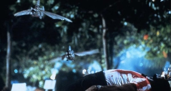 A dead bloody man lies on the ground as piranhas jump over him in Piranha II: The Spawning.
