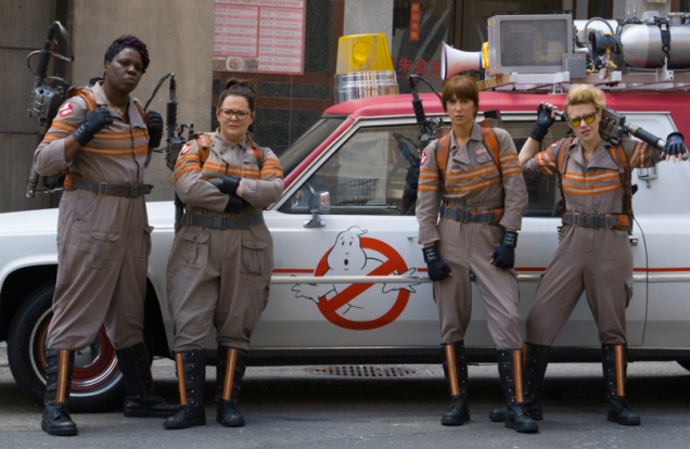 The main cast of the Ghostbusters are standing together in front of their vehicle.