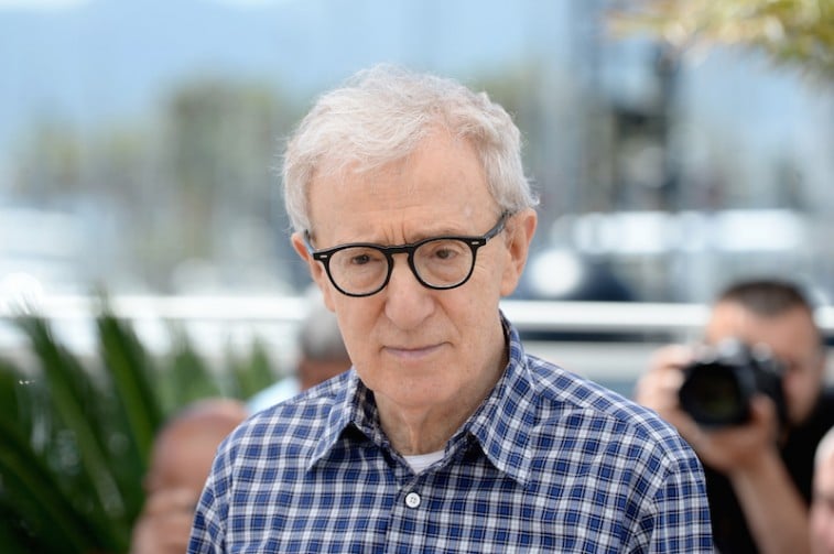 Woody Allen stands in front of the press wearing a flannel shirt and dark glasses.
