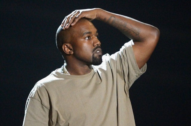 Kanye West in a tan shirt with his hand on his head looking on.