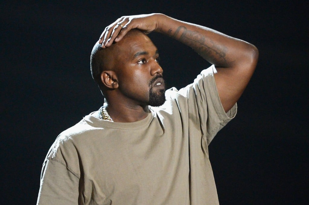 Kanye West in een tan-shirt met zijn hand op zijn hoofd te kijken op