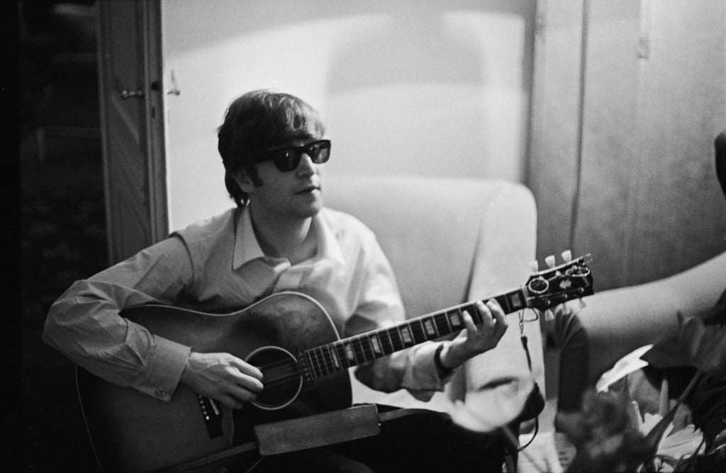 John Lennon playing on an acoustic guitar while sitting on an archair. 