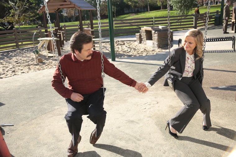 Ron and Leslie hold hands while sitting on swings at a playground. 