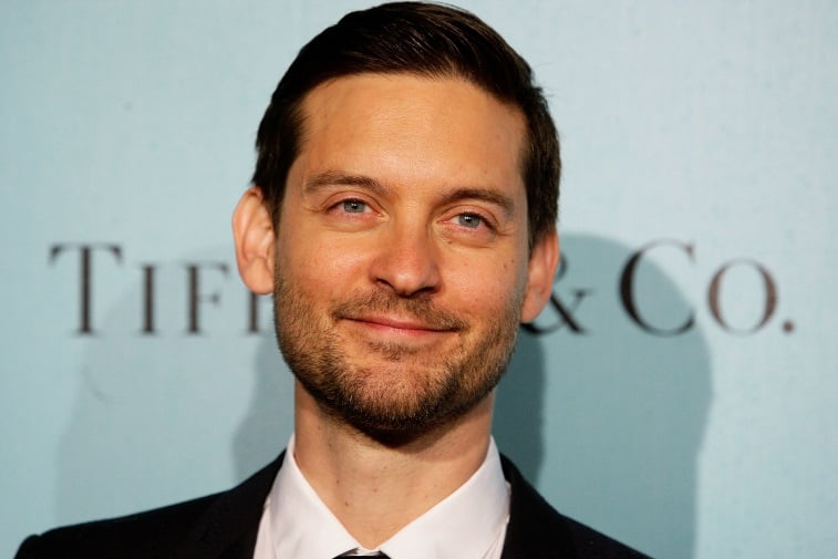 Tobey Maguire smiles while posing in front of a blue wall. 