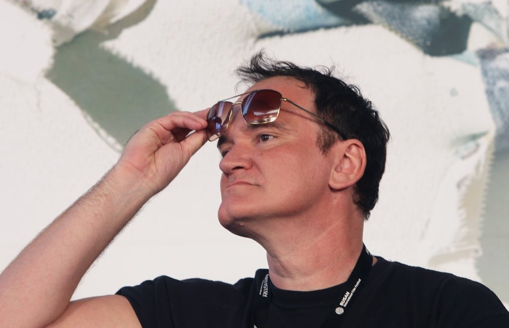 Quentin Tarantino is looking to the side and is lifting his sunglasses from his eyes.