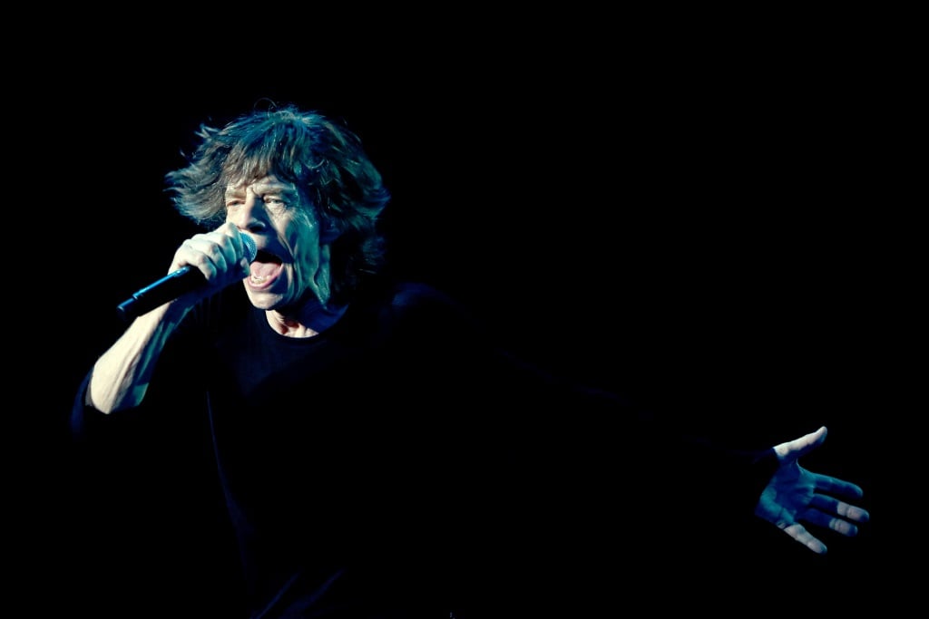 Mick Jagger standing on stage with a microphone near his mouth. Mick Jagger standing on stage with a microphone near his mouth.