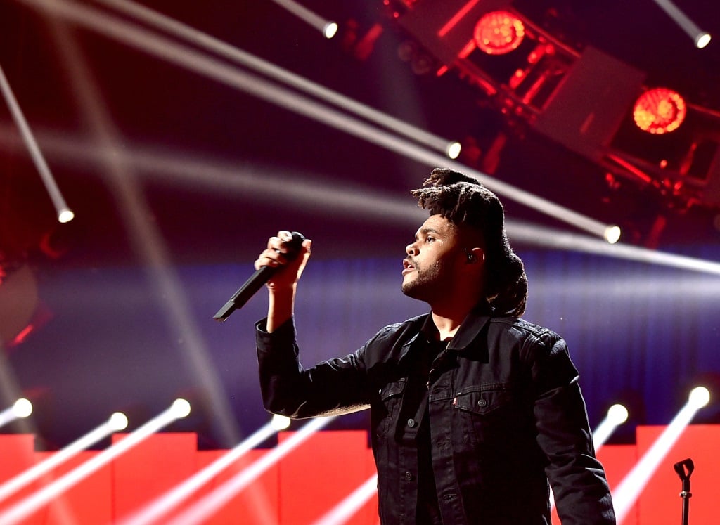 The Weeknd is on stage holding up a microphone and performing on stage.
