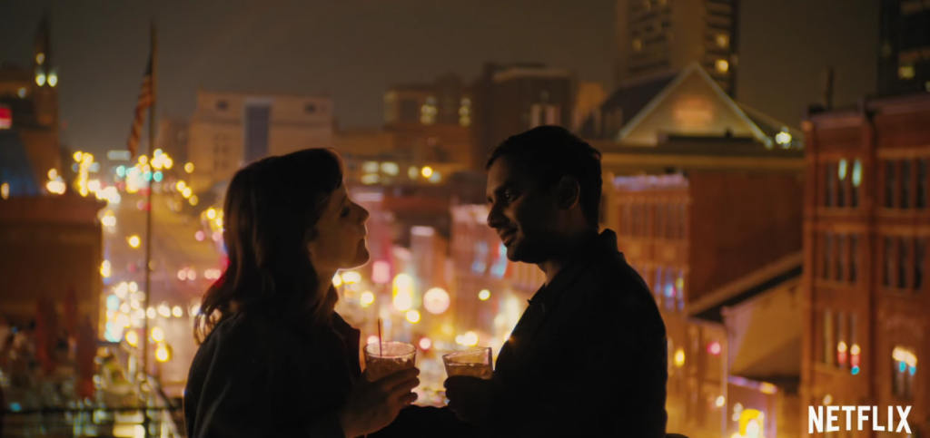 Aziz Ansari and Noël Wells stand on a rooftop at night with drinks in their hands