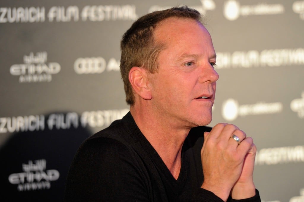 Kiefer Sutherland stares ahead and holds hands together in front of black backdrop that promoting the Zurich Film Festival.