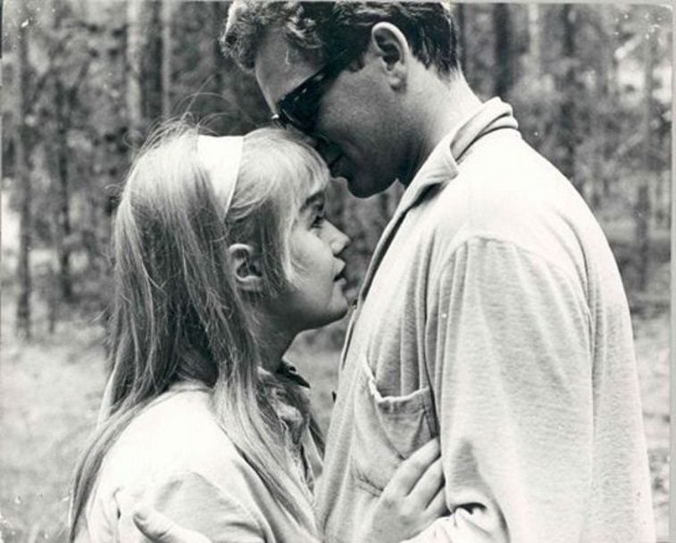 A young woman wearing a headband with her hands on the chest of a man in sunglasses in the woods in I Am Curious (Yellow)