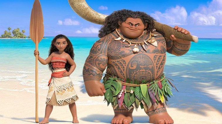Moana and Maui stand next to each other on a beach.