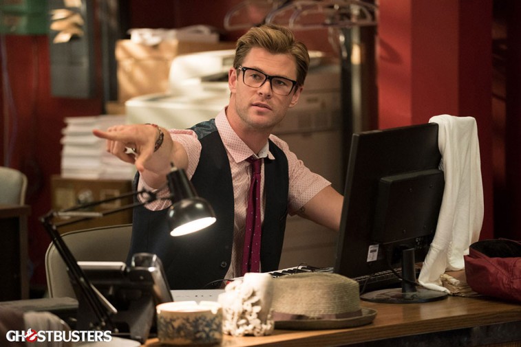 Chris Hemsworth is sitting at a desk and wearing glasses.