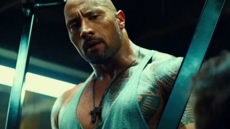 Dwayne Johnson looks down at someone and is wearing a grey muscle shirt in Pain and Gain.