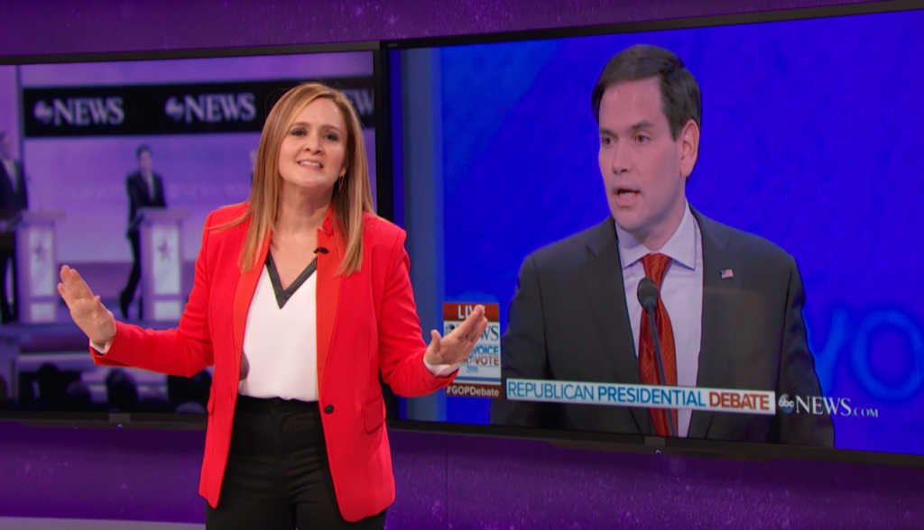 Samantha Bee stands in front of a TV screen