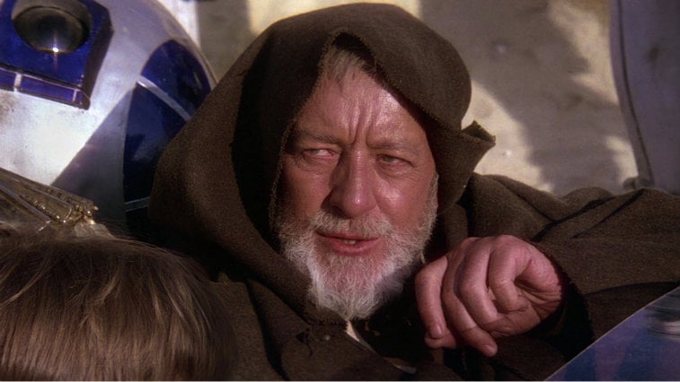 Alec Guinness in a brown robe sitting next to R2-D2.