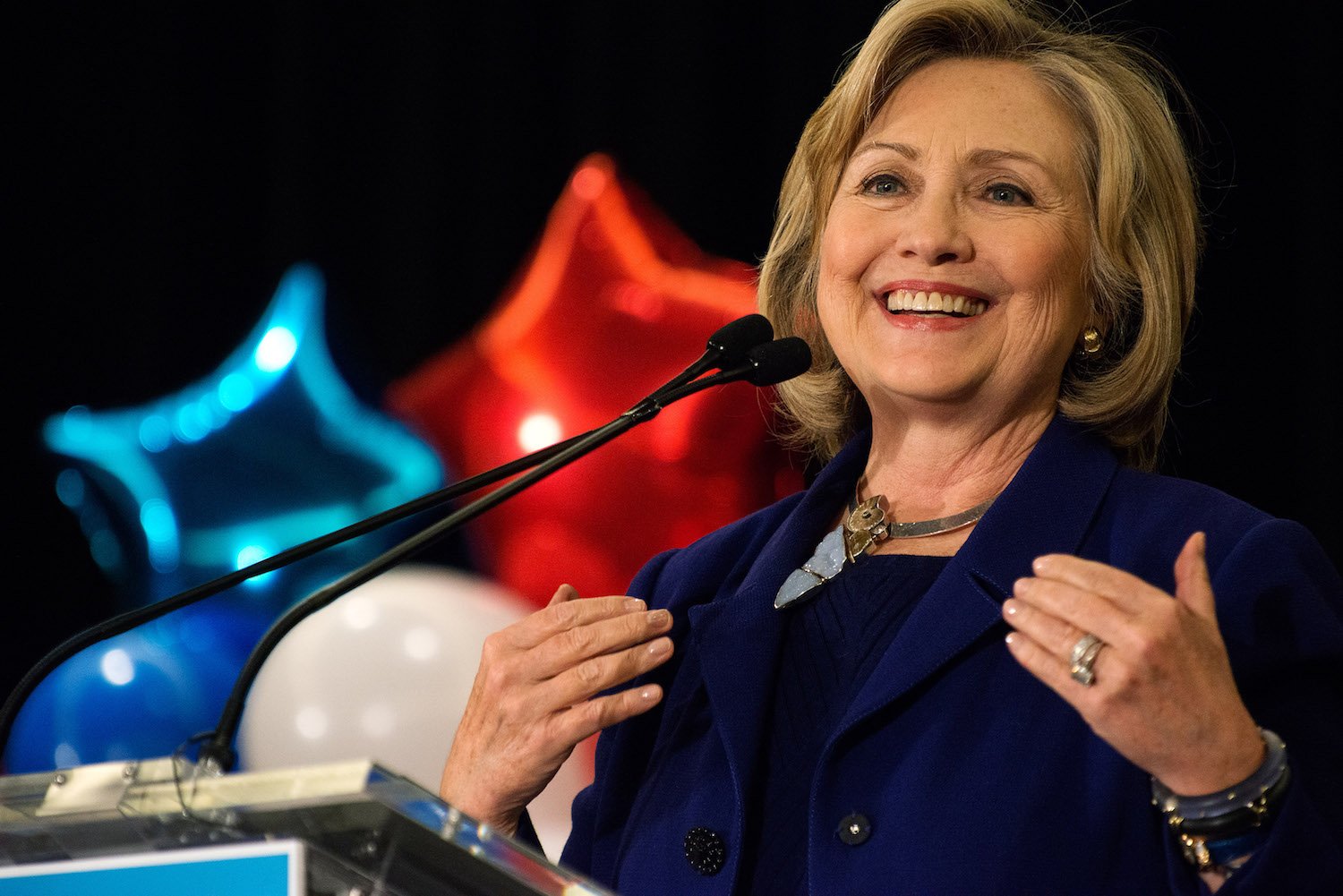 Hillary Clinton giving a speech into a microphone in front of red, white, and blue stars and balloons Hillary Clinton giving a speech into a microphone in front of red, white, and blue stars and balloons