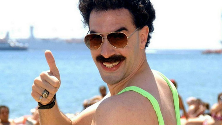 Sacha Baron Cohen posing on a beach in sunglasses and a green swim suit giving a thumbs up in Borat.