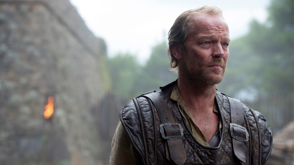 Jorah Mormont is looking ahead and is standing in front of a fortress.