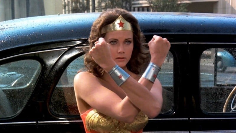 Lynda Carter as Wonder Woman holding her arms in an X-formation in front of a car