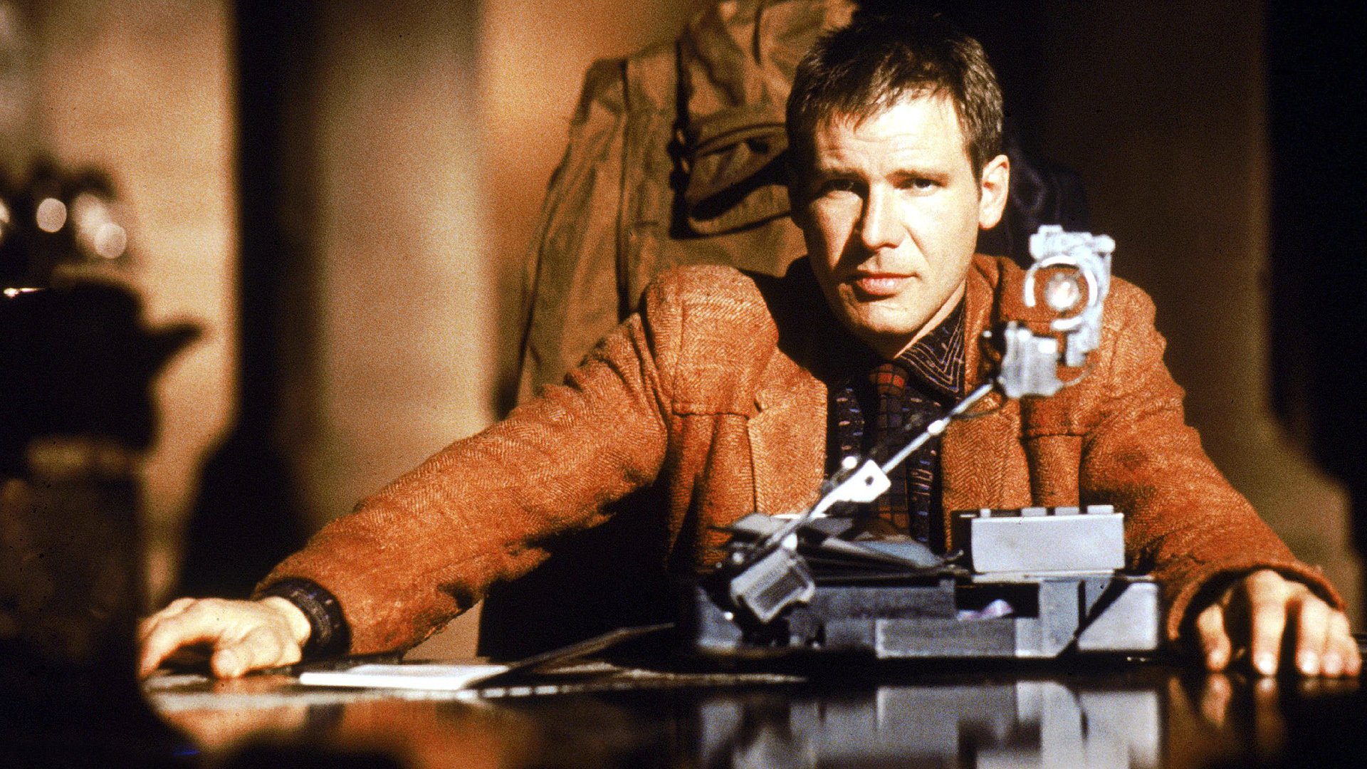 Harrison Ford sits at a desk in front of a machine Harrison Ford sits at a desk in front of a machine