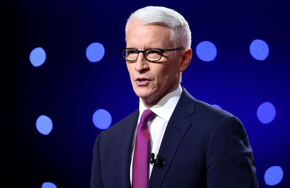 Anderson Cooper stands in a blue suit and purple tie while speaking to an audience.