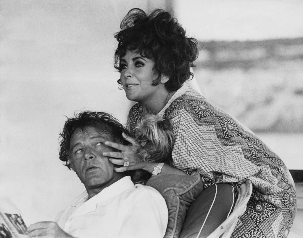 Elizabeth Taylor leans over a chair that Richard Burton is sitting in and touches his face.