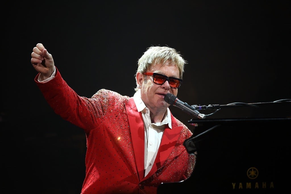 Elton John is in a red suit jacket and playing the piano on stage. Elton John is in a red suit jacket and playing the piano on stage.