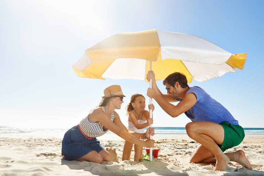 family of three setting up a beach umbrella to limit sun exposure on a sunny day at the beach