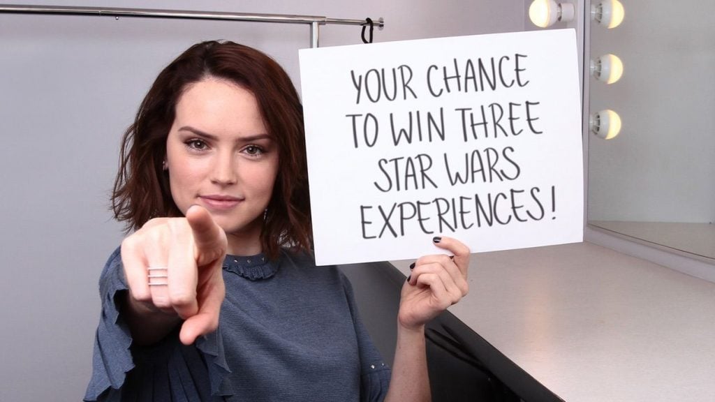Daisy Ridley pointing at the camera, holding up a sign telling fans to enter a giveaway