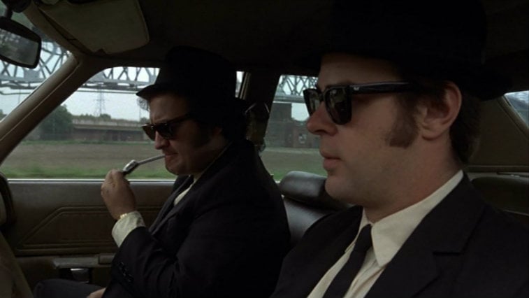 John Belushi and Dan Aykroyd sit in the front seats of a car in suits and sunglasses in The Blues Brothers