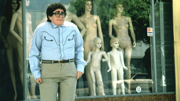 Julia Sweeney stands in front of a storefront window while wearing glasses and shrugging her shoulders in It's Pat