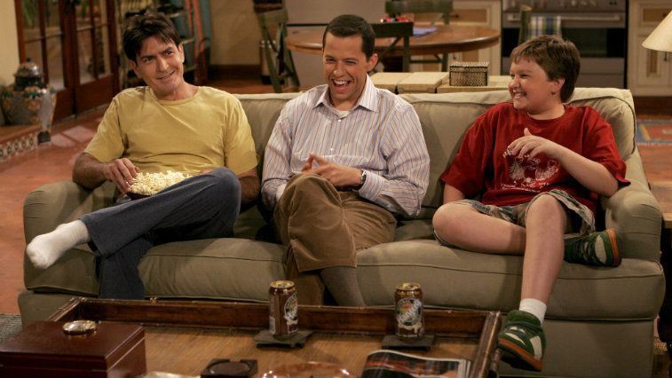 The cast of 'Two and a Half Men' sitting together on a couch as they laugh and watch television. 