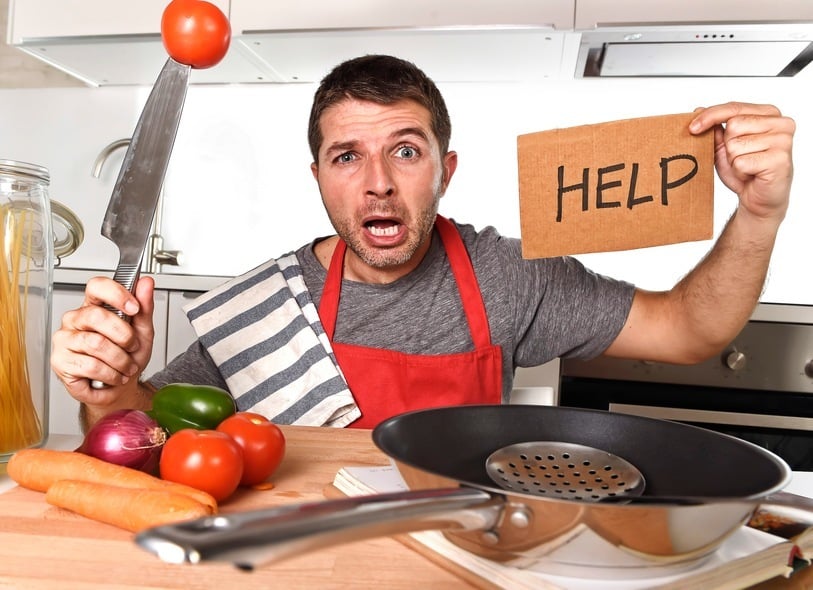 young terrified man at home kitchen wearing cook apron showing help sign looking desperate in stress holding knife with tomato in domestic mess cooking concept