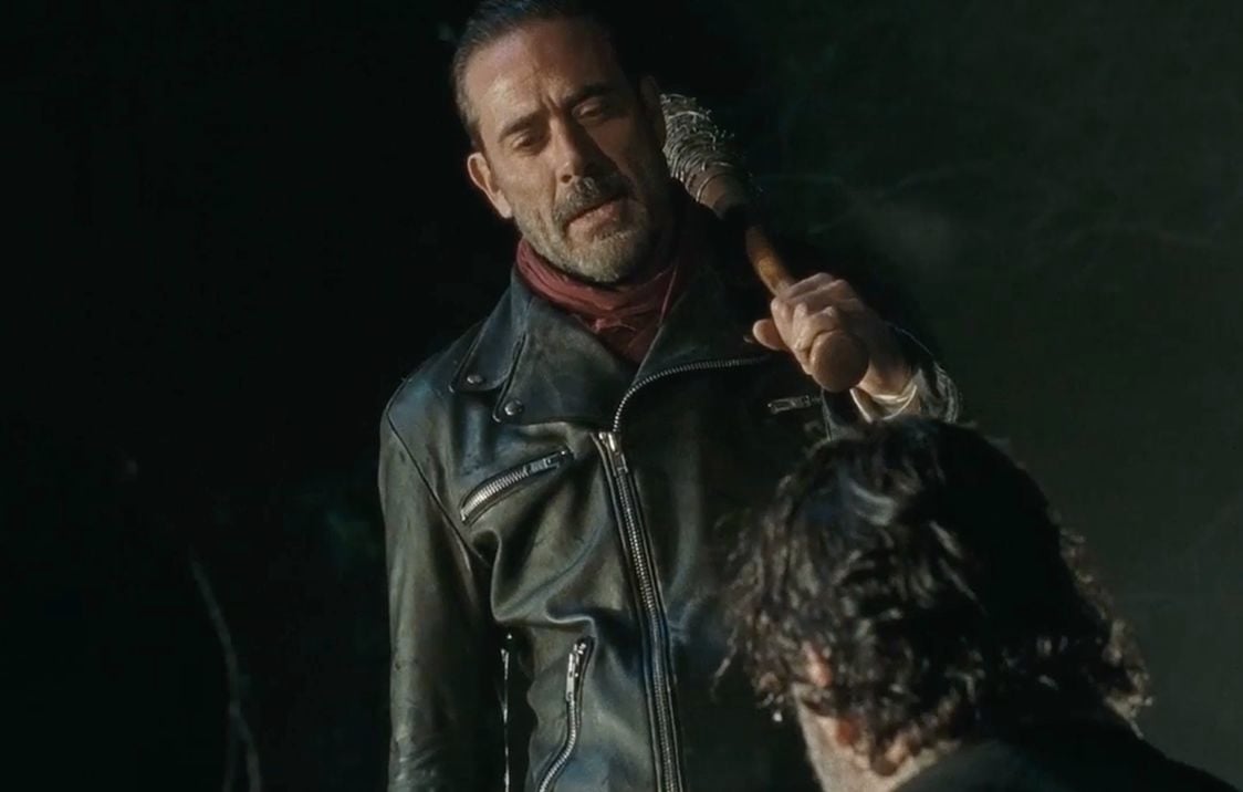 Negan wearing a black leather jacket and red scarf, holding a baseball bat over his shoulder and looking down on a kneeling Rick Grimes