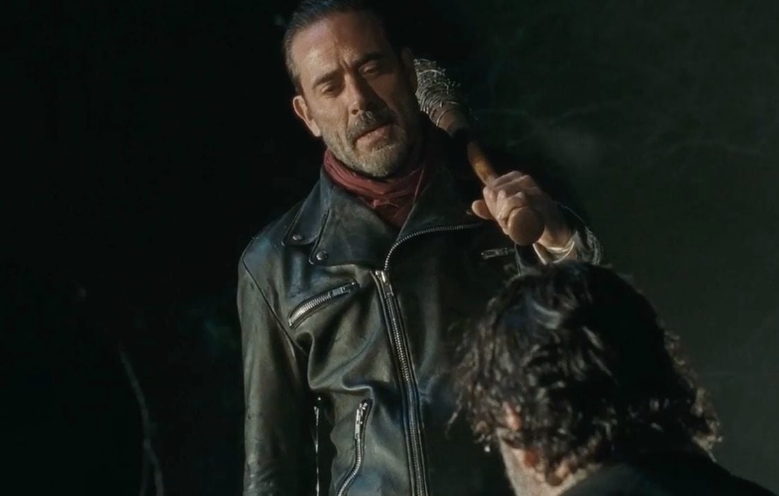 Negan wearing a black leather jacket and red scarf, holding a baseball bat over his shoulder and looking down on a kneeling Rick Grimes Negan wearing a black leather jacket and red scarf, holding a baseball bat over his shoulder and looking down on a kneeling Rick Grimes