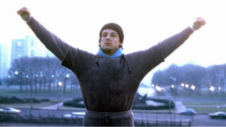 Rocky is at the top of the steps with his arms up in the air.