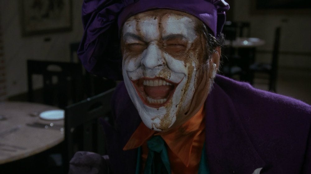 Jack Nicholson as the Joker wearing a purple suit and smiling gleefully while smeared makeup drips down his white face in 'Batman'