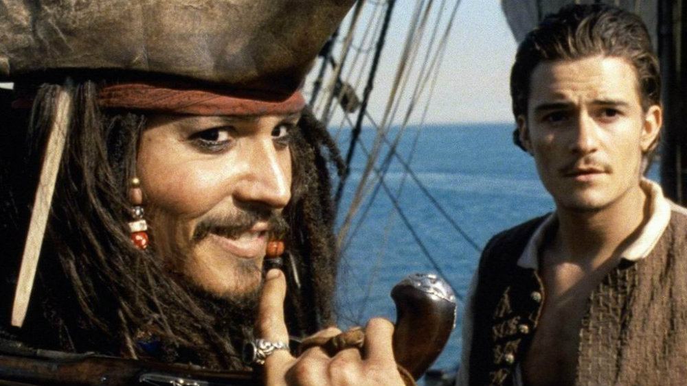 Johnny Depp holding an old gun over his shoulder and smiling, next to a grim-looking Orlando Bloom