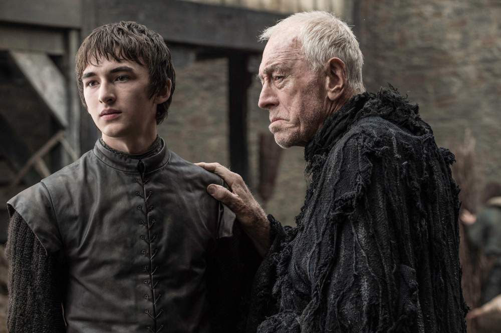 The Three Eyed Raven puts his hand on Bran's shoulder, as they both look off to the left of the frame