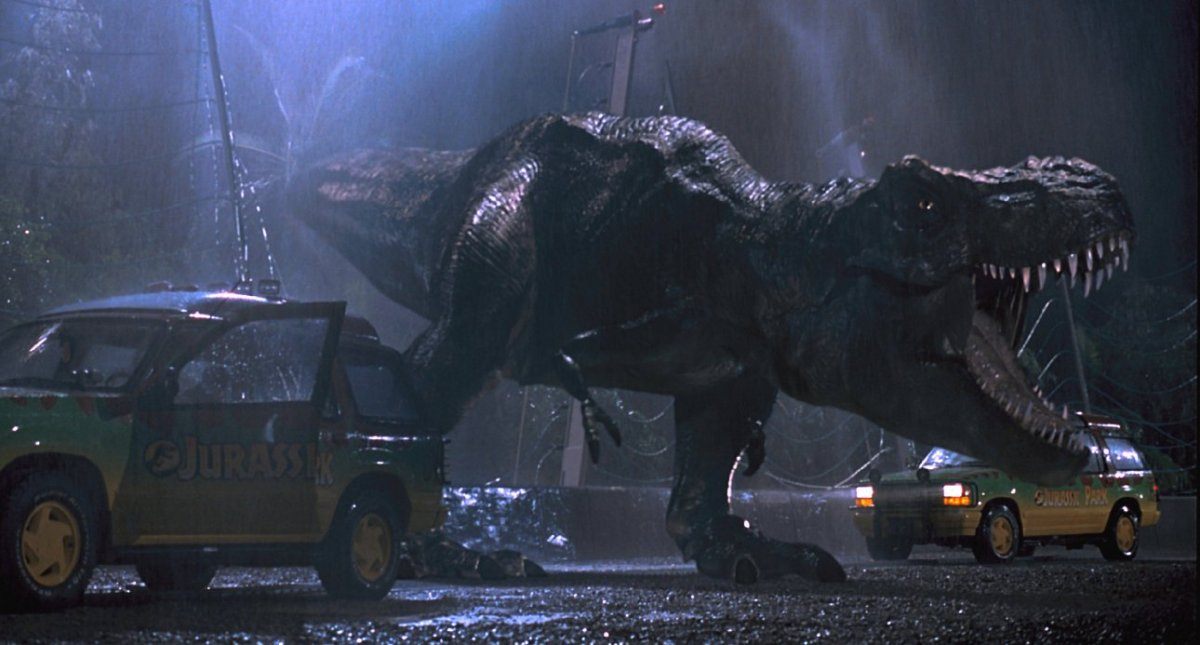 A T-Rex is roaring while standing next to two cars.