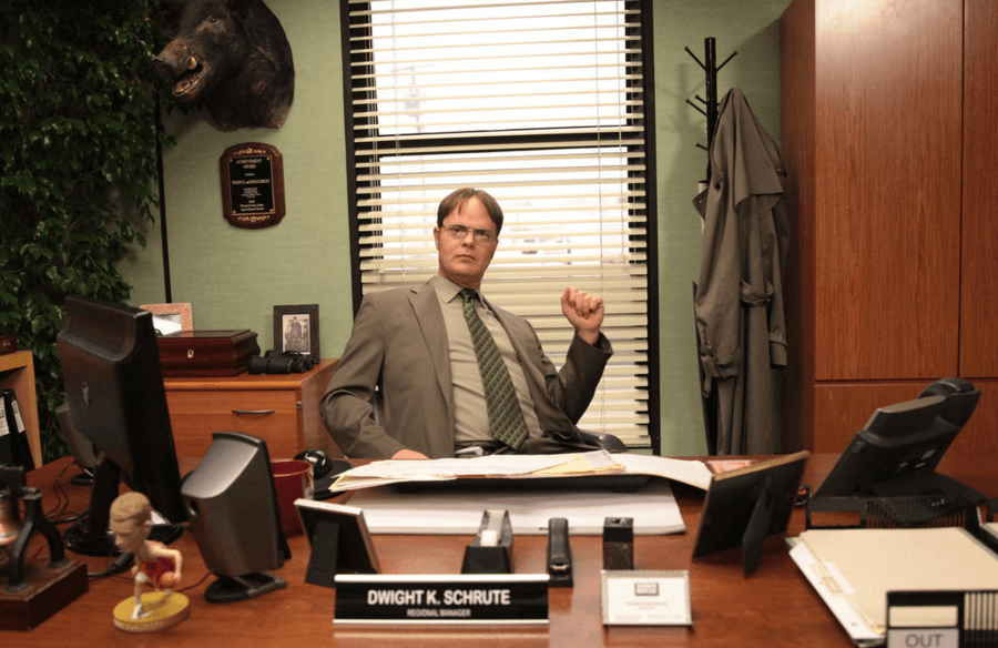 Dwight sits at a desk with supplies laid out neatly in front. 