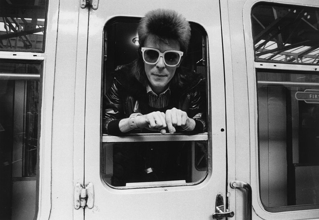 David Bowie at a photoshoot looking out of a train door with sunglasses on