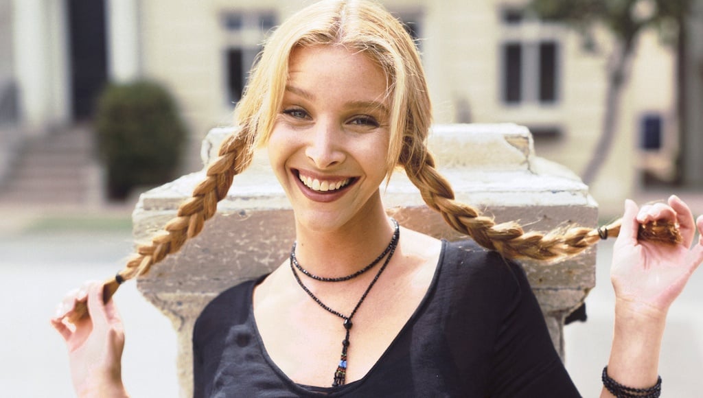 Lisa Kudrow is holding up her braids in each hand and is smiling.