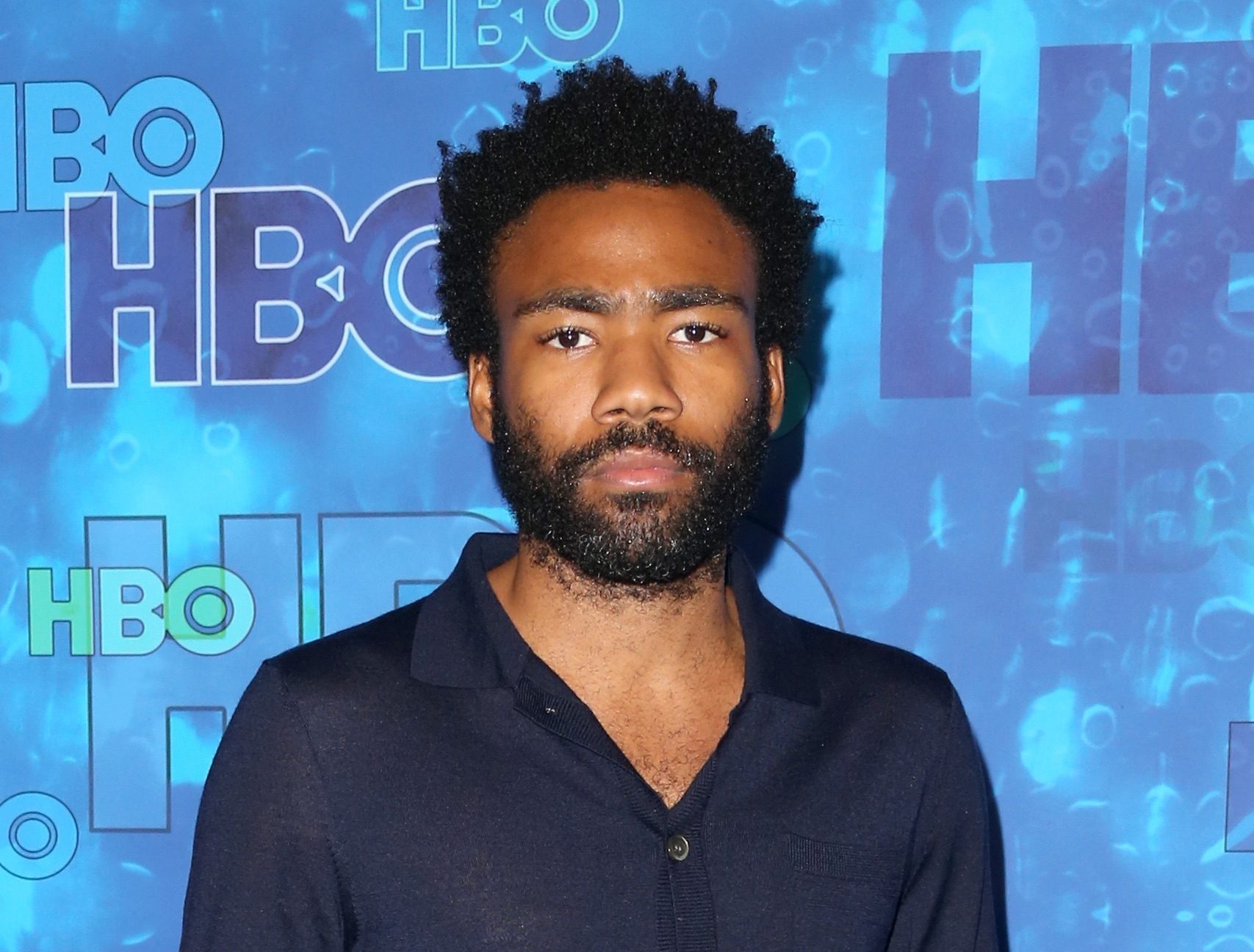 A bearded Donald Glover, looking into the camera in front of an HBO background