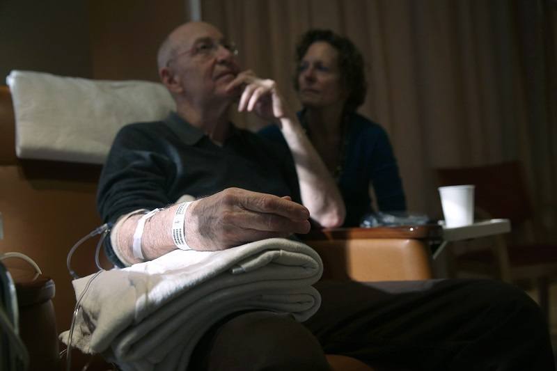 A man sits while undergoing chemotherapy cancer treatment as his wife looks on