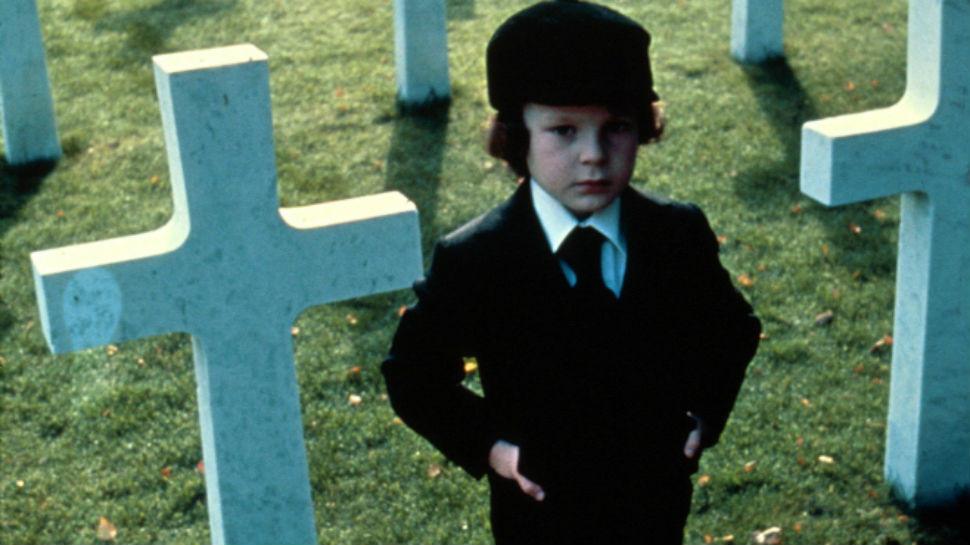 A young boy stands in a graveyard in a black suit and hat in The Omen