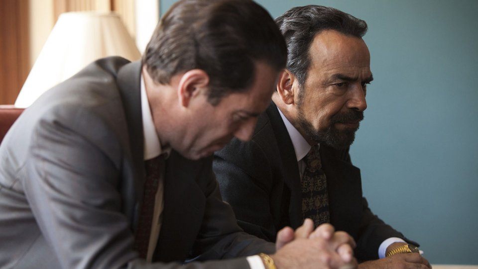 Two men in suits sit next to each other with their hands clasped in Narcos Season 2