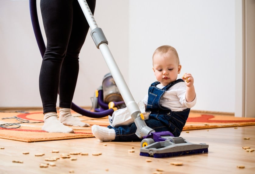 Woman cleaning with vacuum cleaner while baby sitting on floo
