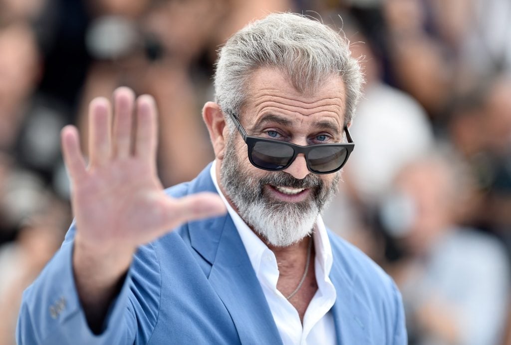 Mel Gibson smiling and waving at the camera, wearing a blue suit and sunglasses.