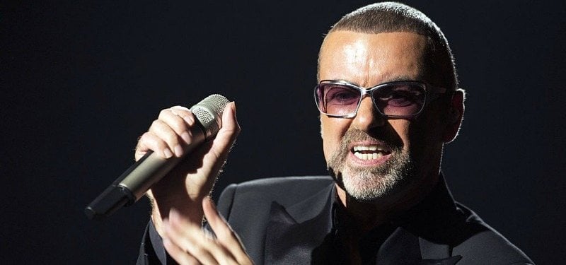 George Michael holding a microphone while wearing a black suit.